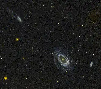 Image d'un ciel étoilé avec plusieurs galaxies visibles. Au centre, une galaxie spirale est bien définie, entourée de quelques autres galaxies plus petites et moins distinctes, dont une en haut à gauche visible depuis la tranche. Plusieurs étoiles brillantes sont également dispersées dans l'image.