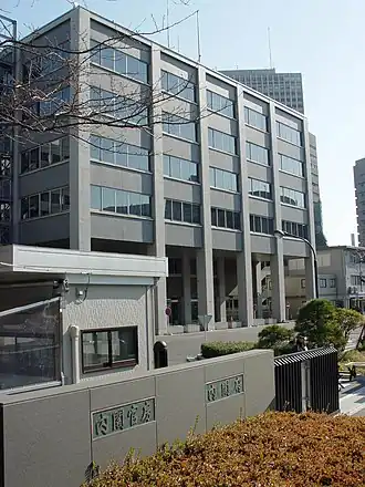 Immeuble moderne en pierre grise de cinq étages, les quatre étages supérieurs s'avançant plus avant, vu de 3/4 gauche. Au premier plan, un mur bas construit avec les mêmes pierres sur lequel est inscrit bureau du cabinet en japonais et un portail noir. À gauche, derrière le mur, se trouve également une cabane de gardien.