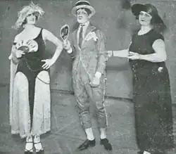 Photographie en noir et blanc. Trois personnes se tiennent debout et ont un chapeau sur la tête : un homme est au milieu et à gauche et à droite se trouvent deux femmes. Les deux femmes portent une robe. La robe à gauche de l'image et bicolore blanc/noir, la robe à droite est noire. L'homme est vêtu d'un complet gris.