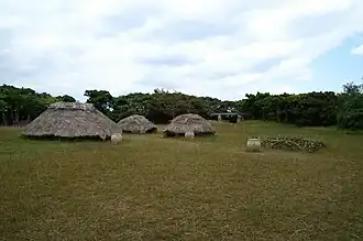 Photographie du village de la période Kaizuka du site de Nakabaru (reconstruit). Elle montre trois huttes avec un toit fait de branchages dans une clairière couverte d’herbe, et une autre hutte, effondrée, qui permet de voir la structure interne des habitations.