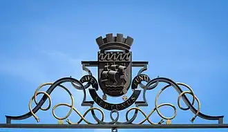 Écu sur fond de ciel bleu, sur un support de fer forgé aux formes courbes.