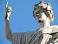 Haut d'une statue blanche, on voit la tête et un bras, un doigt pointant vers le ciel.