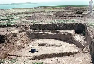 Chantier archéologique dans lequel on peut voir une excavation d'une vingtaine de metres carré de deux à trois metres de profondeur. La terre est beige. On voit la base d'une maison ronde. Plus loin on apperçoit d'autre trous similaires sans en voir le contenu. À l'horizon, on distingue une étendue d'eau.