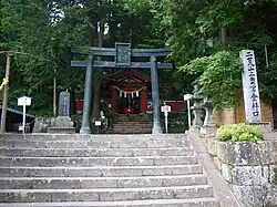 Photo couleur d'un torii en haut d'un escalier en pierre. Des arbres aux feuilles vertes forment l'arrière-plan.