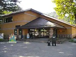 Photo couleur de l'entrée d'un bâtiment aux portes vitrées, et aux murs en briques oranges. Un ciel blanc laiteux en arrière-plan, et un sol pavé gris au premier plan.