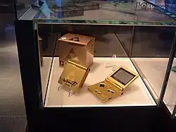 Sous une vitrine, on observe deux consoles de jeu dorées et logotées.