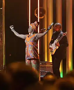 chanteuse africaine levant les bras au ciel en tenant un micro dans sa main gauche, à l'arrière plan un guitariste.