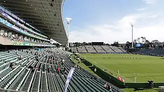 vue d'un stade depuis une tribune avec au centre de l'image la pelouse et au fond une autre tribune non couverte
