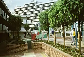 À l'avant-plan, une petite place avec un muret en pierre décorée. Quelqu'un se promène dans une allée bordée d'arbres. En fond, un grand immeuble et quelques canalisations surélevées.