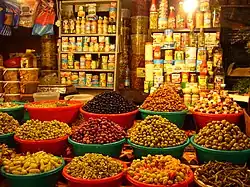 photo en couleurs, au premier plan des bassines en plastique remplies d'olives et de piments, au fond des étagères portent des boites de conserve et des bouteilles d'huile