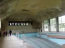Une piscine abandonnée au toit incurvé. Des grandes fenêtres font la longueur de la piscine, et des plus petites sont au fond. Trois personnes non identifiables se tiennent debout sur le rebord de la piscine.