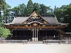 Photo couleur de la façade ombragée de la salle du culte d'un sanctuaire shintō, structure en bois foncé dont le frontispice est doré par endroits. L'arrière-plan est constitué d'une rangée d'arbres au feuillage vert sous un ciel bleu sans nuages.