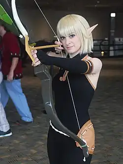 Cosplay d'une femme, en habit moulant noir, cheveux blonds et oreilles pointues. Elle bande un grand arc, prête à tirer.