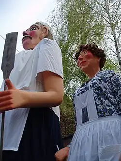 Les géants de Oudezeele : Julia la souriante cantinière se trouve derrière Jeannot le boulanger. Cheveux frisés, pommettes rouges saillantes, nez pointu surmonté de lunettes en ailes de papillon, Julia porte une blouse à fond blanc et motifs de feuillages et fleurs bleues, un collier de perles vertes et un tablier blanc à bavette, garni de dentelle blanche.