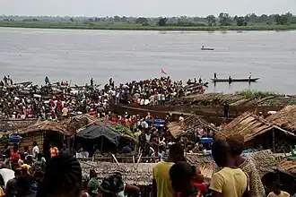 Photographie couleur montrant une foule et des pirogues au bord d'un large fleuve.
