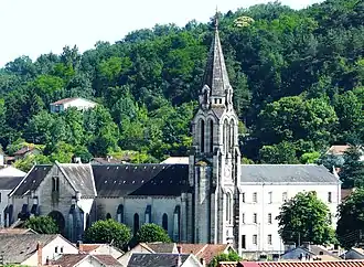 Photo représentant sur fond de verdure l'église Saint-Georges. Au centre, son clocher flèche et sur la gauche sa nef de laquelle ressort son transept proéminent. À droite du clocher, un long et haut bâtiment de trois étages situé derrière l'église donne l'impression d'une autre nef, presque symétrique.