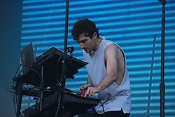 Homme caucasien portant un débardeur blanc face plusieurs consoles de mixage.