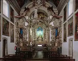 Photographie en couleurs. Intérieur de la nef : les murs sont blancs et affublés de nombreux tableaux ; les angles sont forces de reliefs en couleurs, très baroques, tandis que l'autel au fond semble illuminé d'une lueur verte.