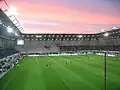 Photo de l'intérieur du stade depuis une tribune derrière le but. On distingue des joueurs sur la pelouse, des tribunes remplies et un parcage quasi vide, avec un ciel rosé par le soleil couchant. Les hautes parois et auvents isolent le stade de l'extérieur.