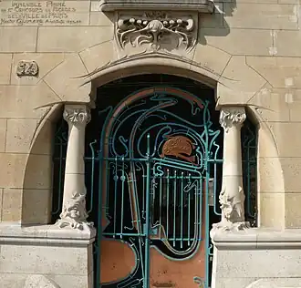 Photo en couleur d'une base de façade claire percée d'un arc, avec deux colonnettes sculptées et un portail en métal bicolore tout en arabesques