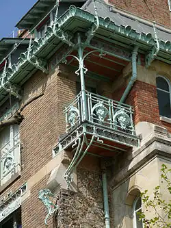 Photo en couleur d'un coin d'immeuble avec matériaux de diverses couleurs, et ornements de balcon et de gouttière en fer forgé clair, bleu-vert