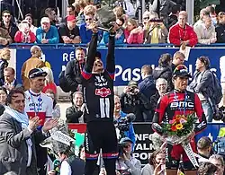 Photographie en couleur représentant, au premier plan, devant la foule, au centre le vainqueur levant le trophée, un pavé, au dessus de sa tête, encadré par les deuxième et troisième portant un bouquet de fleur