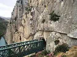 Sur la gauche de l'image un pont ferroviaire vert et sur la droite une falaise avec une passerelle où des piétons circulent