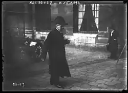 Paul Bourget en tenue d'académicien, habit, manteau et bicorne, moustache, tient un document dans la main gauche, peut-être le discours de réception d'Émile Boutroux. Il marche en regardant le sol pavé de la cour d'honneur de l'Institut de France, quai de Conti. Une automobile est garée à gauche de la photographie sur verre. Un homme, au second plan, chapeau et manteau, adossé à la façade du bâtiment, près de deux fenêtres avec barreaux, observe l'académicien en portant sa main gauche au visage et en tenant un sac dans sa main droite. Une hampe en bois (?) disparaît dans l'angle droit du négatif sur verre.