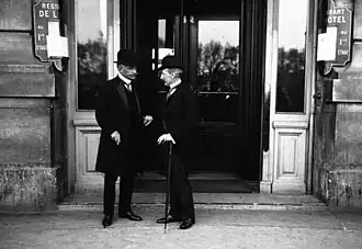 Photo en noir et blanc de deux hommes en train de converser ; l’homme à gauche, moustachu et cheveux gris-blancs, porte un chapeau, une longue veste, un veston, une cravate et une chemise à blanche à faux col
