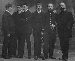 Photo en noir et blanc de six hommes se tenant debout. Parmi eux, quatre jeunes hommes portent chacun un complet ; un homme plus âgé arbore une tenue militaire, avec un homme plus vieux, barbu, s'appuyant sur son épaule