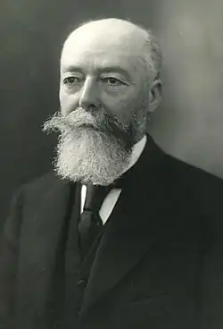 Portrait photographique d'un homme aux cheveux et à la barbe grisonnants, vêtu d’une chemise blanche, d’un veston, d'une veste et d'une cravate foncés