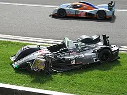 Photographie d'une voiture de sport-prototype verte, blanche, noire et rouge, vue de haut et de trois-quarts, sur de la pelouse, et d'une voiture de sport-prototype bleu et orange vue de profil
