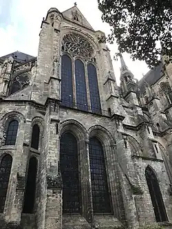 Petit transept, en partie caché par un arbre, avec sa rosace en forme de fleurs et les deux grandes verrières de la partie basse. À droite, bâtiment de brique.