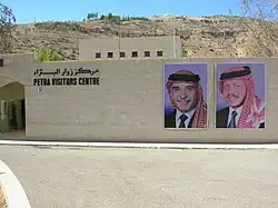 Photographie d'un mur moderne pourvu d'une enseigne en anglais et en arabe. Sur la gauche du mur, il y a une entrée vitrée et, sur la droite, deux affiches représentent chacune le visage d'un homme différent. Ils sont tous deux coiffés d'un keffieh. Une rue passe devant le mur.