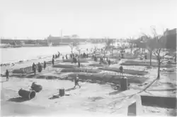 Photo noir et blanc montrant plusieurs fosses récemment rebouchées, avec des gens déambulant autour, au bord d'une rivière.