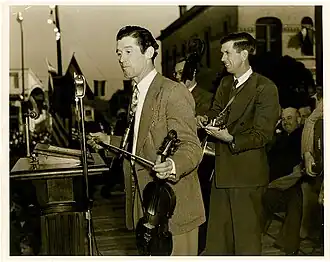 un homme en veste debout sur une scène devant un micro, avec un violon dans une main et un archet dans l'autre, musiciens derrière lui