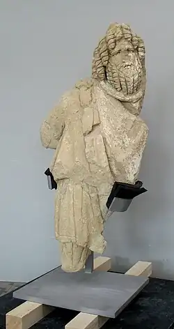 Vue d'une statue en pierre d'un personnage barbu, sans bras ni jambes, coiffé d'un bonnet phrygien.