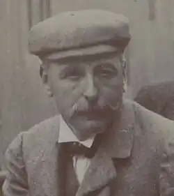 Photo en portrait d'un homme arborant une moustache en brosse et un couvre-chef.