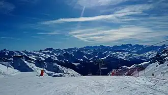 Photographie de pistes de ski, damées ; en face on voit les montagnes bleues enneigées.
