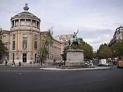Photographie en couleur d'une place ornée d'une statue de cavalier, à gauche un immeuble en rotonde, à droite une avenue