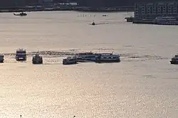 Photo prise au moment du coucher du soleil. L'avion est au centre de l'image, dans le fleuve tendant vers le jaune-orange dû à la réflexion du soleil couchant. Sur l'image, au moins sept ferries entourent l'avion ainsi qu'un hélicoptère en haut à gauche de l'image.
