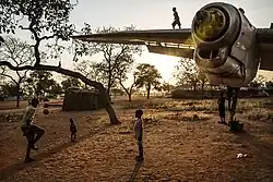 Photographie en couleurs d'enfants jouant autour et sur la carcasse d'un avion, dont on ne voit qu'une aile. L'image est prise au coucher du Soleil.