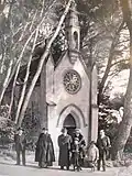 Plusieurs personnes dont un prêtre catholique en habit liturgique devant la chapelle du Plantier, entourée de grands pins.