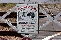 Gros plan sur le panneau émaillé du portillon qui porte l'indication : « NE TRAVERSEZ PAS sans regarder dans les DEUX DIRECTIONS. Un train peut en CACHER un autre » et un dessin avec une famille qui traverse alors que survient une locomotive à vapeur qui leur était masquée par un wagon.