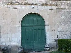 photo couleur montrant une porte en bois à deux battants peinte en vert délavé. Le linteau en arc de cercle est surmonté par une corniche supportée par les pilastres qui encadrent la porte.