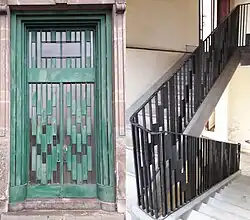 Photographie en couleurs représentant à gauche une porte d’entrée en ferronnerie verte et à droite un escalier blanc avec une main courante en ferronnerie noire.