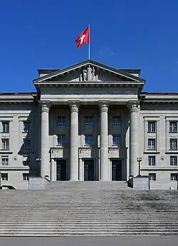 Photographie d'un monument, au premier plan un grand escalier, au centre quatre grandes colonnes encadrant les portes d'entrée, le toit en v comprend une statue en son centre et est surmonté d'un drapeau rouge à croix blanche