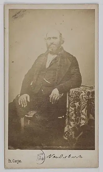 Photographie en noir et blanc représentant un homme mûr barbu en costume trois pièces sombre assis de trois-quart face sur un fauteuil et accoudé à un guéridon recouvert d'un tissu.