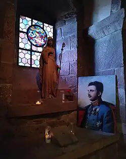 Photographie d'un vitrail devant lequel se trouvent une statue en pierre d'une femme et un peinture représentant le portrait un homme.