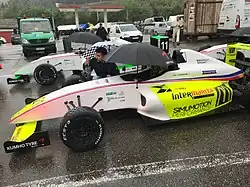 Photographie d'une monoplace de F4 blanche et jaune fluo, vue de profil gauche, devant une autre voiture, à l'arrêt, sur une piste humide.